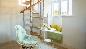 A spiral staircase with metal railings and wooden steps, a fluffy chair, a small round table with a vase of flowers, a green sofa with yellow cushions, and a window with a view of greenery. - Stunning contemporary cottage - The Bothy (Bibury)