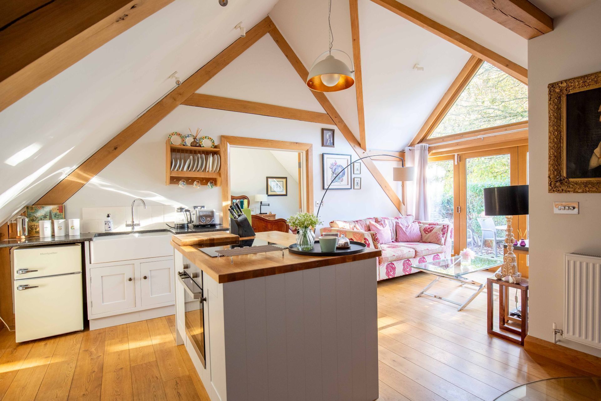 A kitchen with a central island, wooden beams, a dining area with a pink sofa, and a view of the outdoors.
