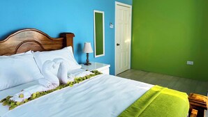 Un lit bien fait, avec des draps blancs et un plaid vert, une tête de lit en bois, une table de chevet avec une lampe et un miroir accroché au mur.