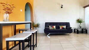 Un salon moderne avec un canapé noir, une table basse en verre et un coin repas comprenant une table blanche et des bancs.