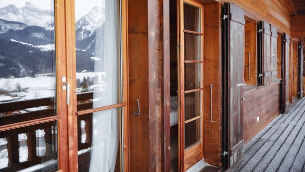 Une cabane en bois dotée de grandes fenêtres et de grandes portes, d’une terrasse en bois et offrant une vue sur des montagnes enneigées.