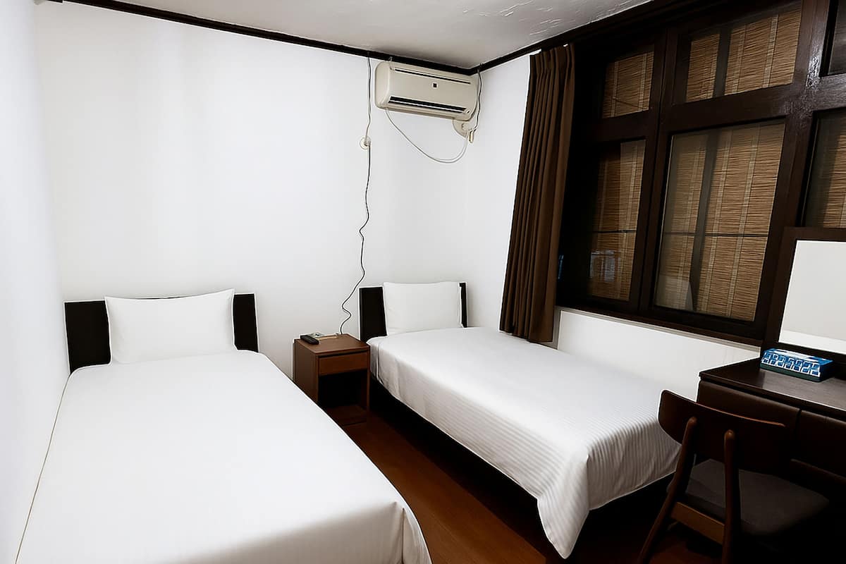 A hotel room with two single beds, a desk with a chair, a window with curtains, and an air conditioning unit.