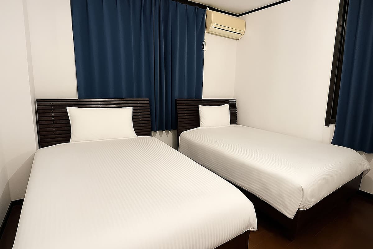 A hotel room with two single beds, a dark wooden headboard, white linens, and a ceiling air conditioner.