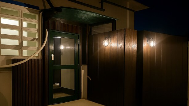 A hallway with a green door, a white wall with a window, and a wooden wall with two light fixtures. - NAGACHIBI (Miyakojima)