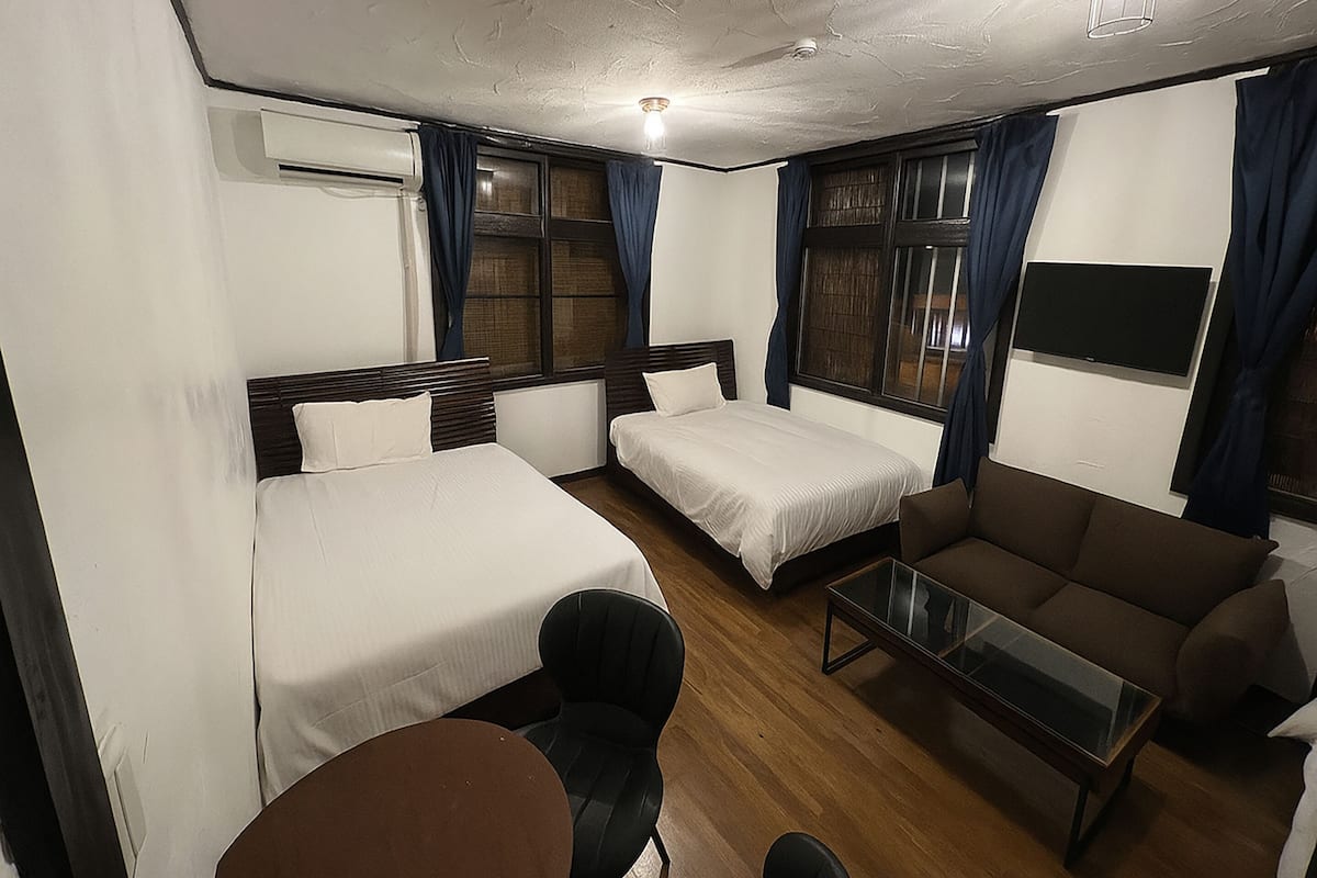 A hotel room with two single beds, a sofa, a small table, and an air conditioning unit.