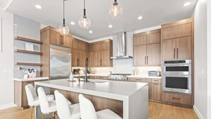 Una cocina moderna con una isla grande, electrodomésticos de acero inoxidable, armarios de madera e iluminación colgante.