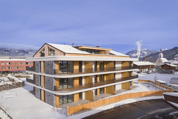 Un immeuble moderne à plusieurs étages, doté de balcons, qui se dresse sur fond de montagnes enneigées.