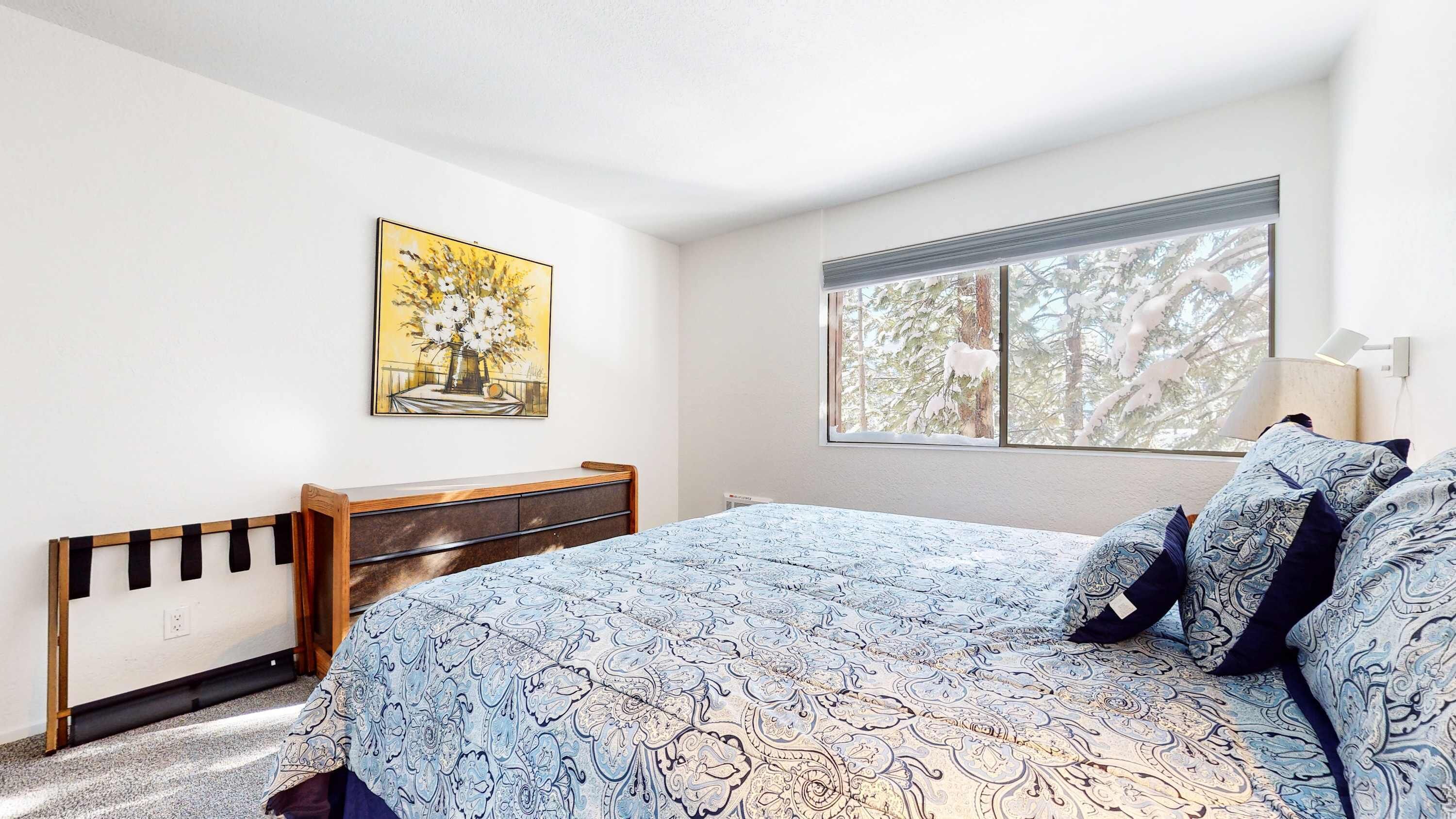 A bedroom with a bed, a bedside table, a lamp, a window with a view of trees, and a painting on the wall.