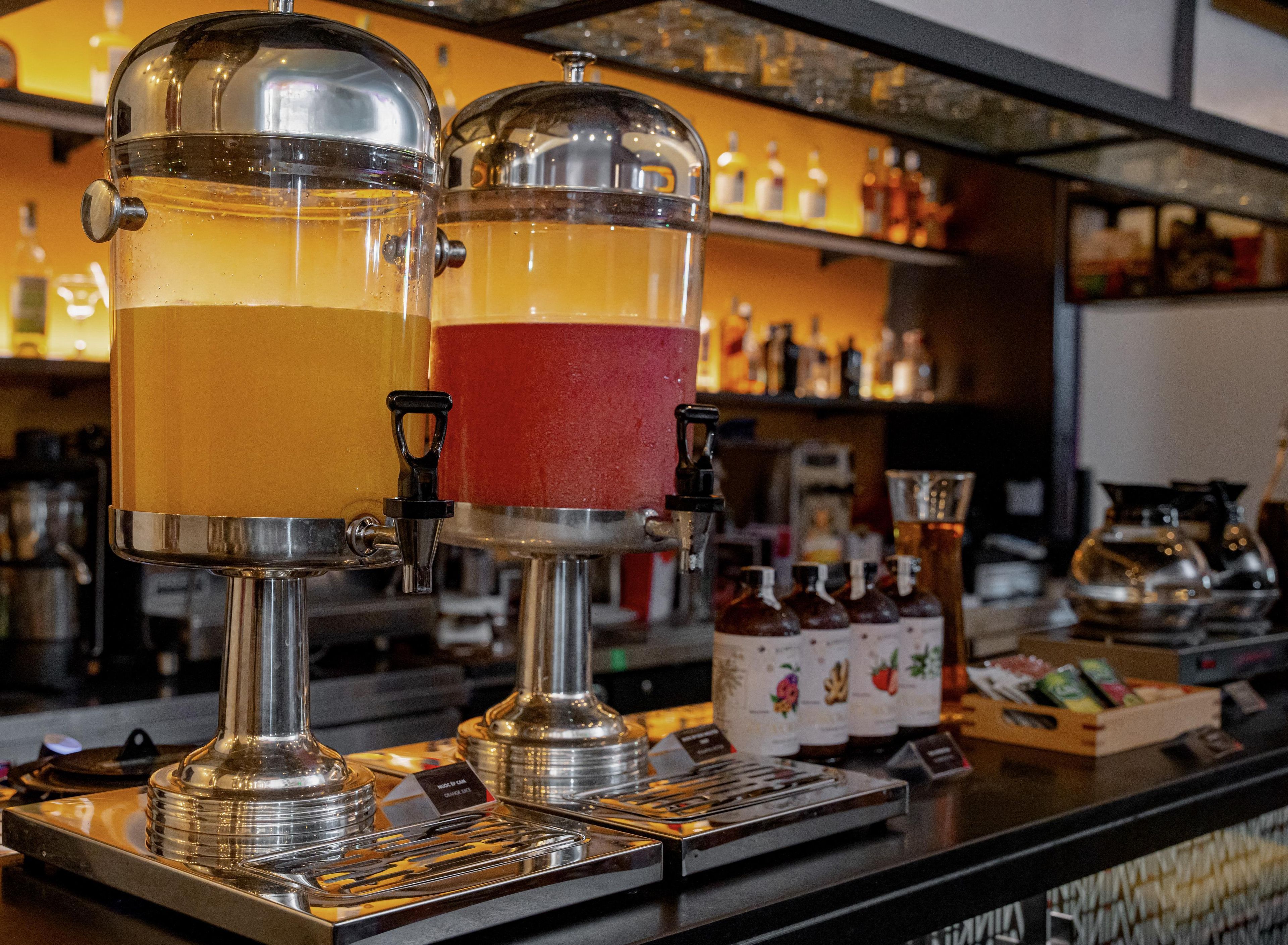 Dois dosadores de bebidas com bicos e torneiras, um balcão de bar e prateleiras com garrafas ao fundo.