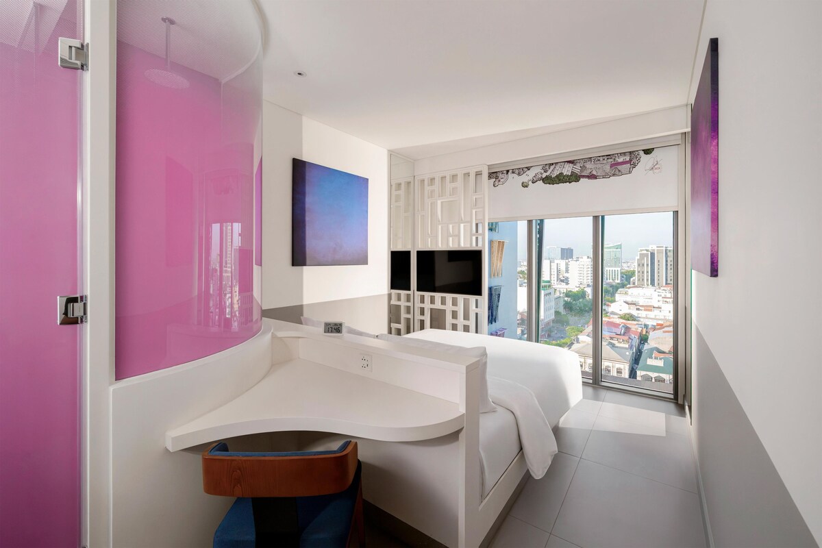 Une salle de bain moderne avec un lavabo aux lignes courbes, un grand miroir, un téléviseur à écran plat et une vue sur la ville par une grande fenêtre.