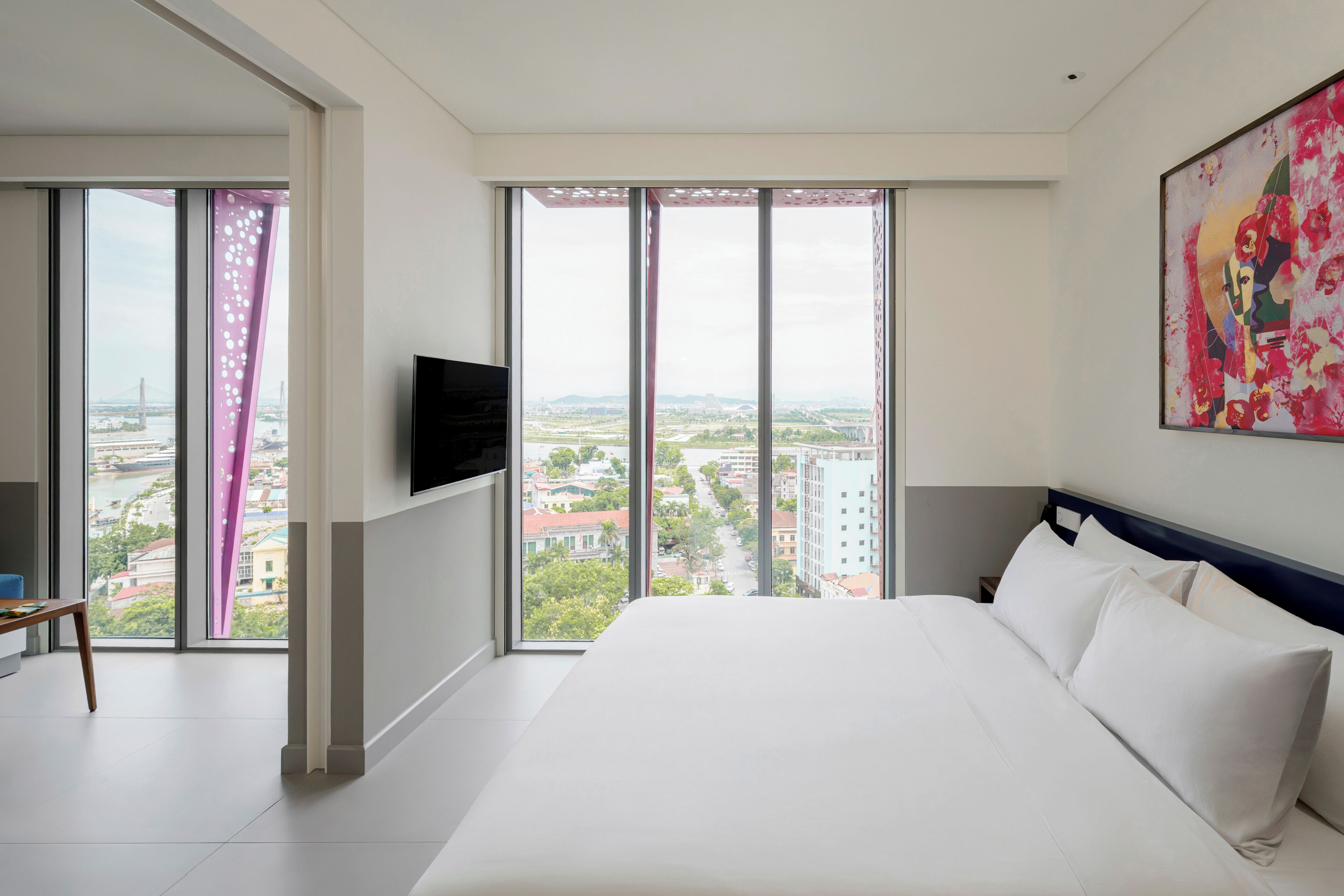 Habitación de hotel con una cama grande, un televisor de pantalla plana montado en la pared y vistas a la ciudad a través de ventanales de gran altura.