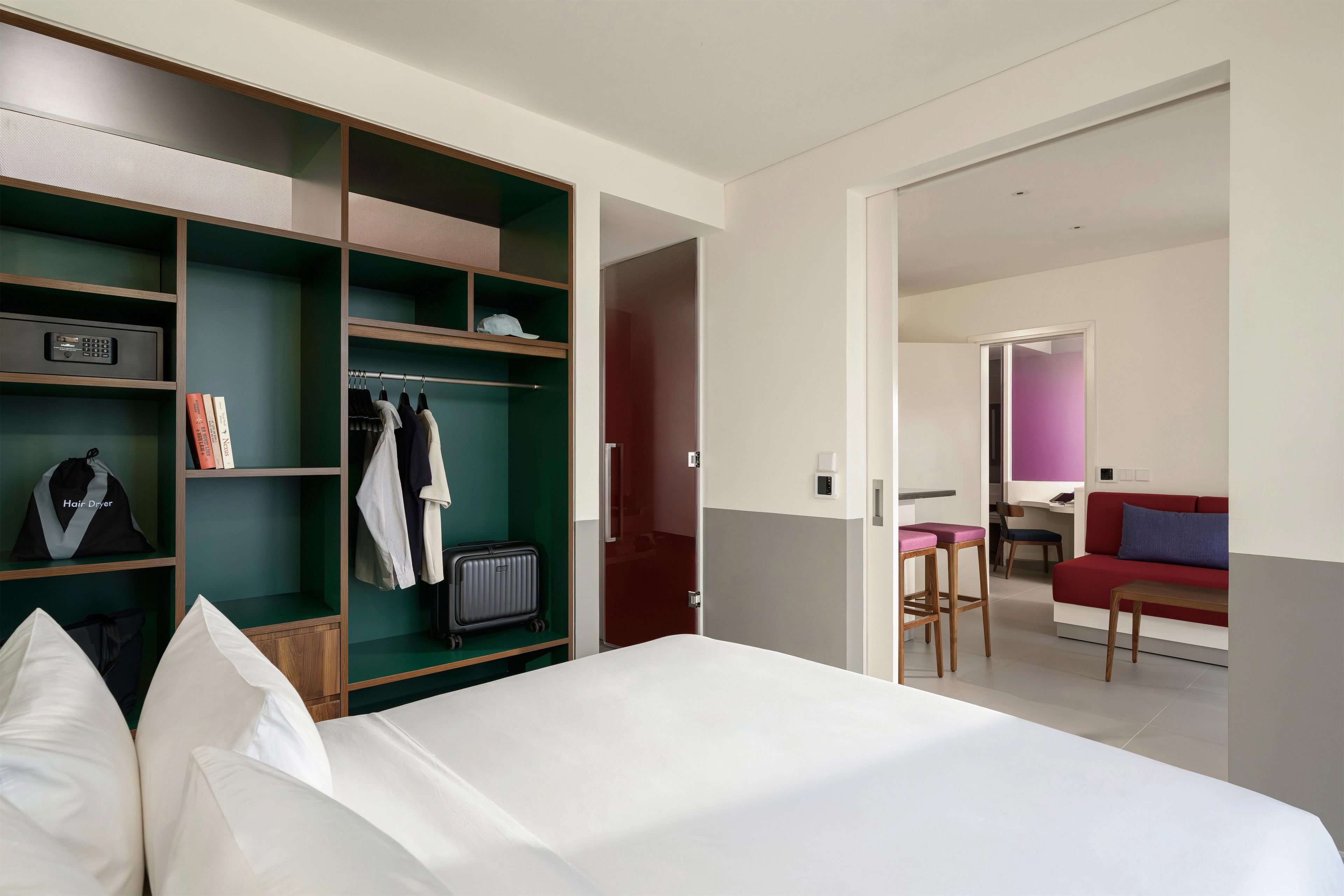 A hotel room with a bed, a wardrobe with hanging clothes, a suitcase, and a hair dryer on the shelf.