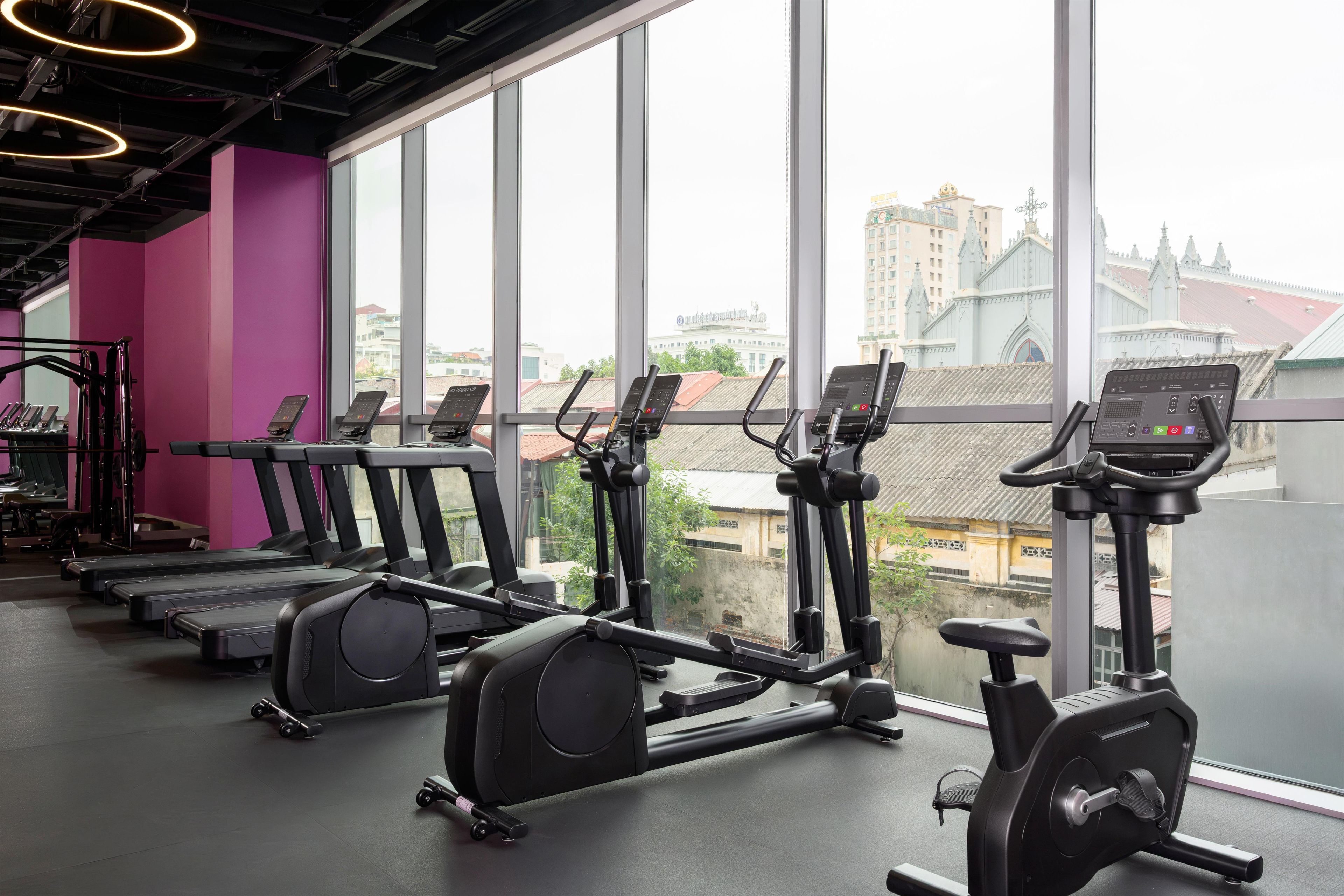 Une salle de sport équipée de tapis de course, de vélos elliptiques et d’un rameur, avec de grandes fenêtres donnant sur des bâtiments et un pont.