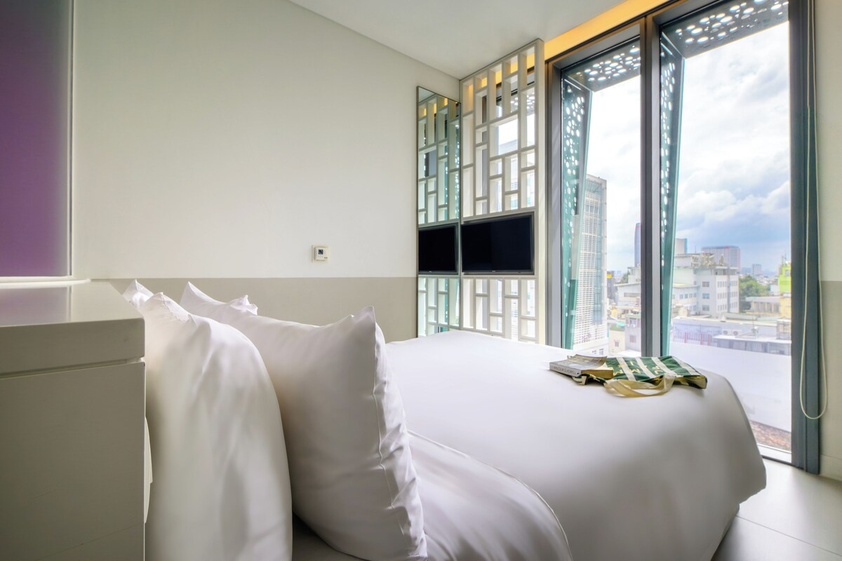 A hotel room with a bed, pillows, a window with a view of buildings, and a door with a decorative screen.