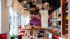 Ein modernes Restaurant-Interieur mit Holztischen und bunten Hocker, Pendelleuchten und einer violetten Akzentwand.