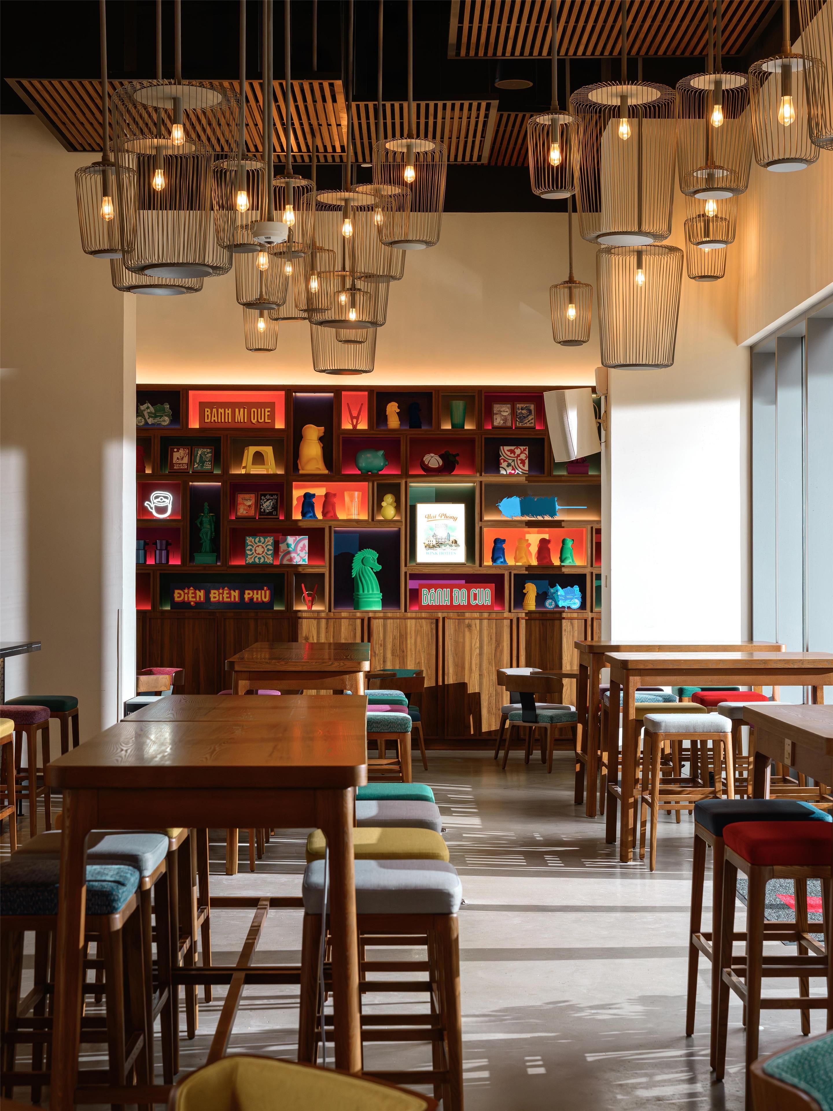 A restaurant interior with wooden tables and chairs, hanging pendant lights, and a wall display of various items.