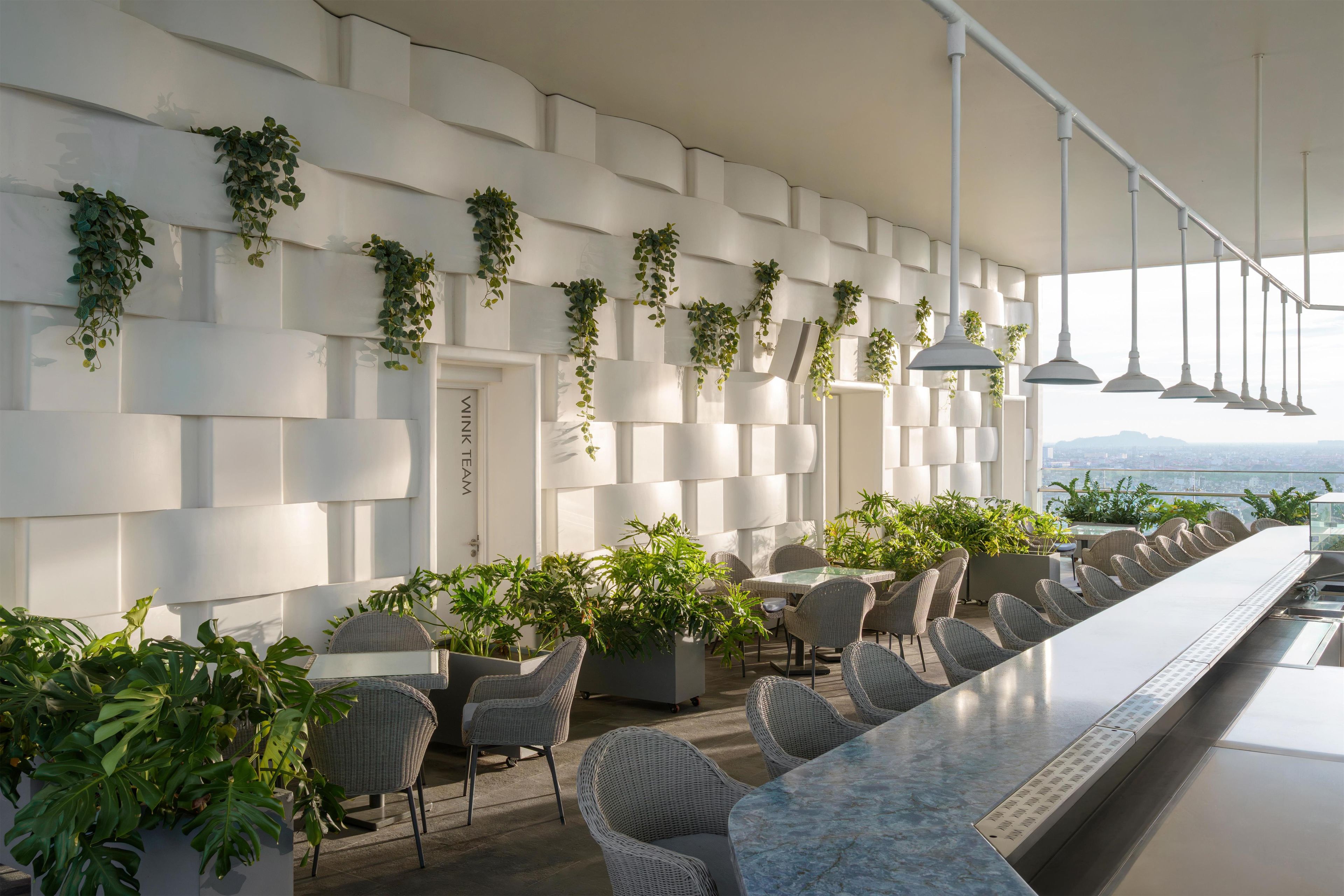 A modern bar area with white walls, hanging plants, wicker chairs, and a long counter.