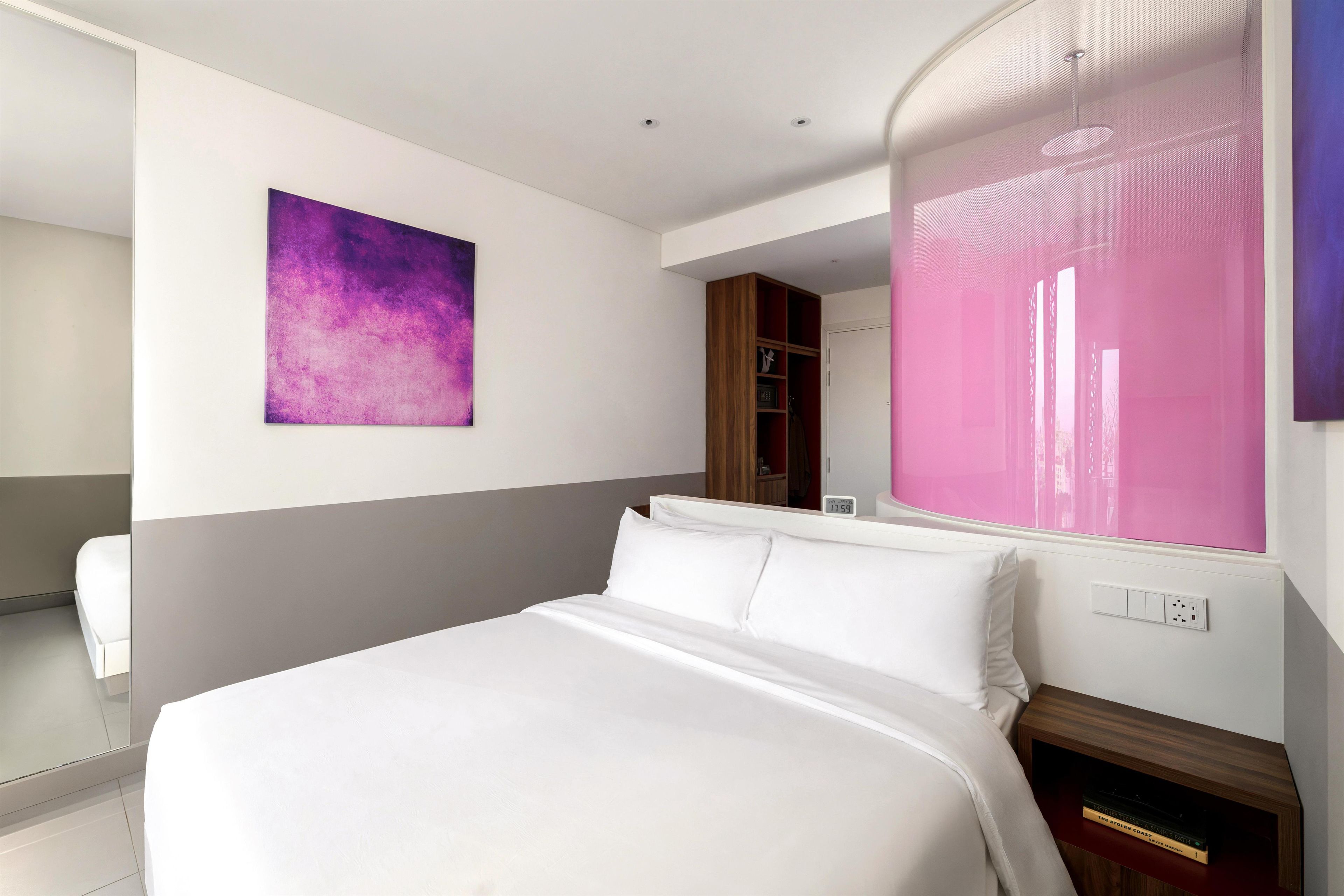 A modern hotel room with a large bed, a painting on the wall, a wardrobe, and a window with a view.