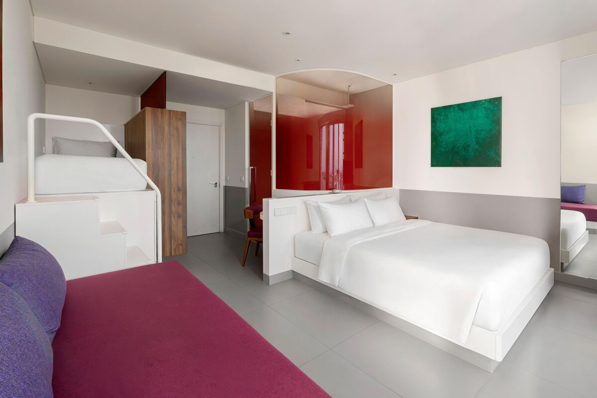 Une chambre d’hôtel avec un grand lit, un bureau et une chaise. Une porte donne accès à une autre pièce.