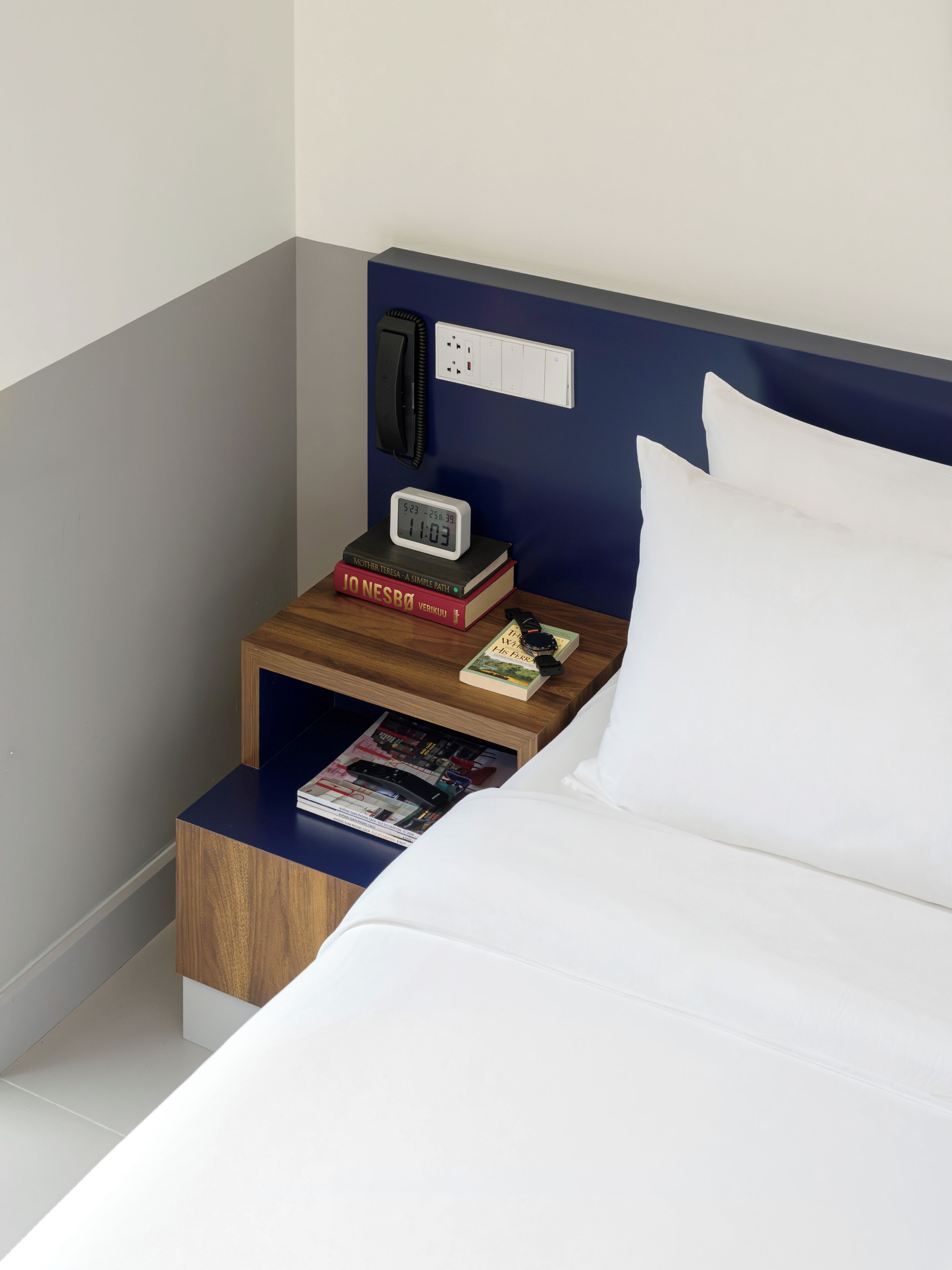 Un lit bien fait, avec des draps blancs, une table de chevet sur laquelle on peut voir une horloge, des livres et un magazine.