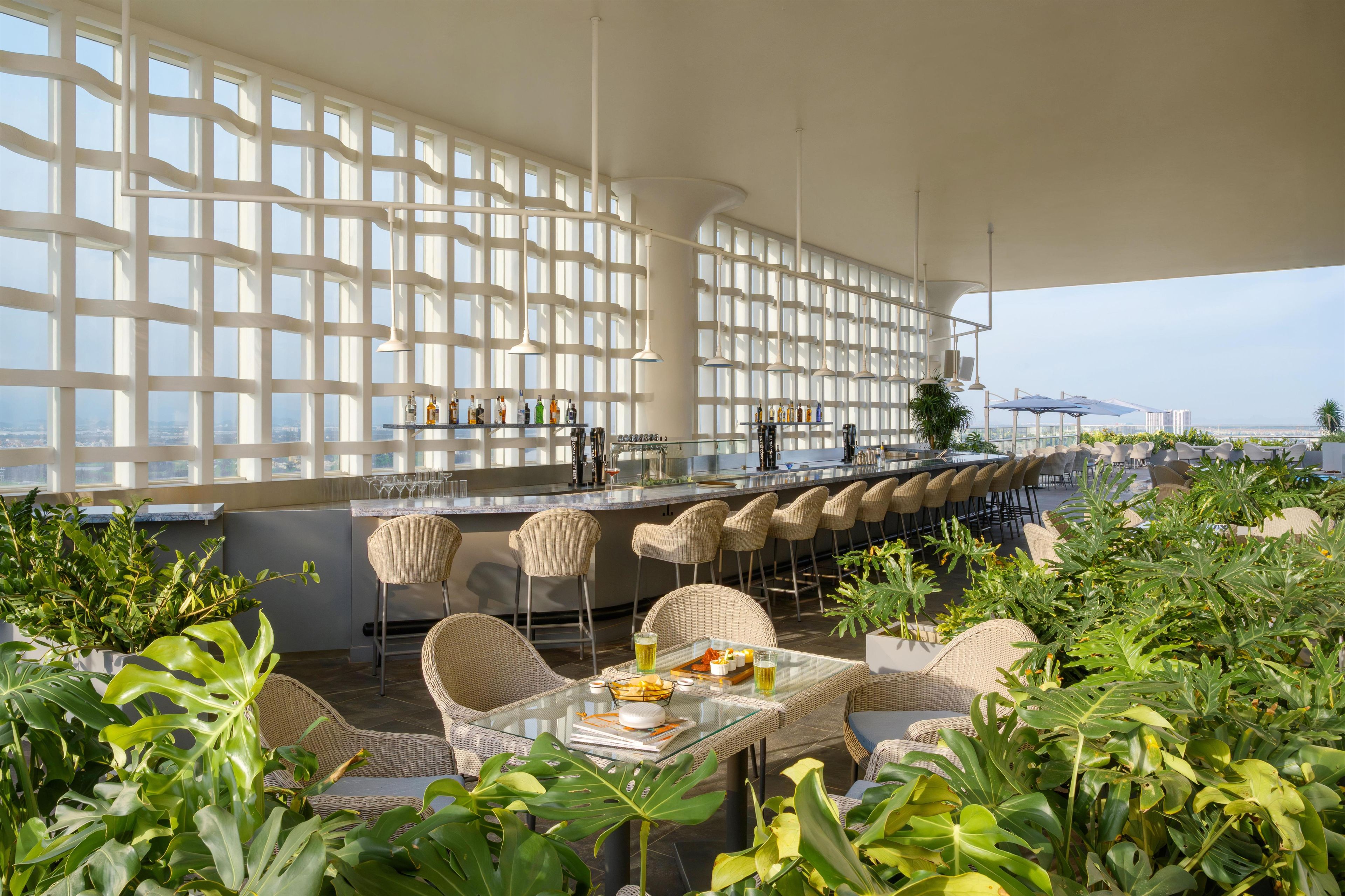 A rooftop bar with a long counter, wicker chairs, and a glass table with a drink.