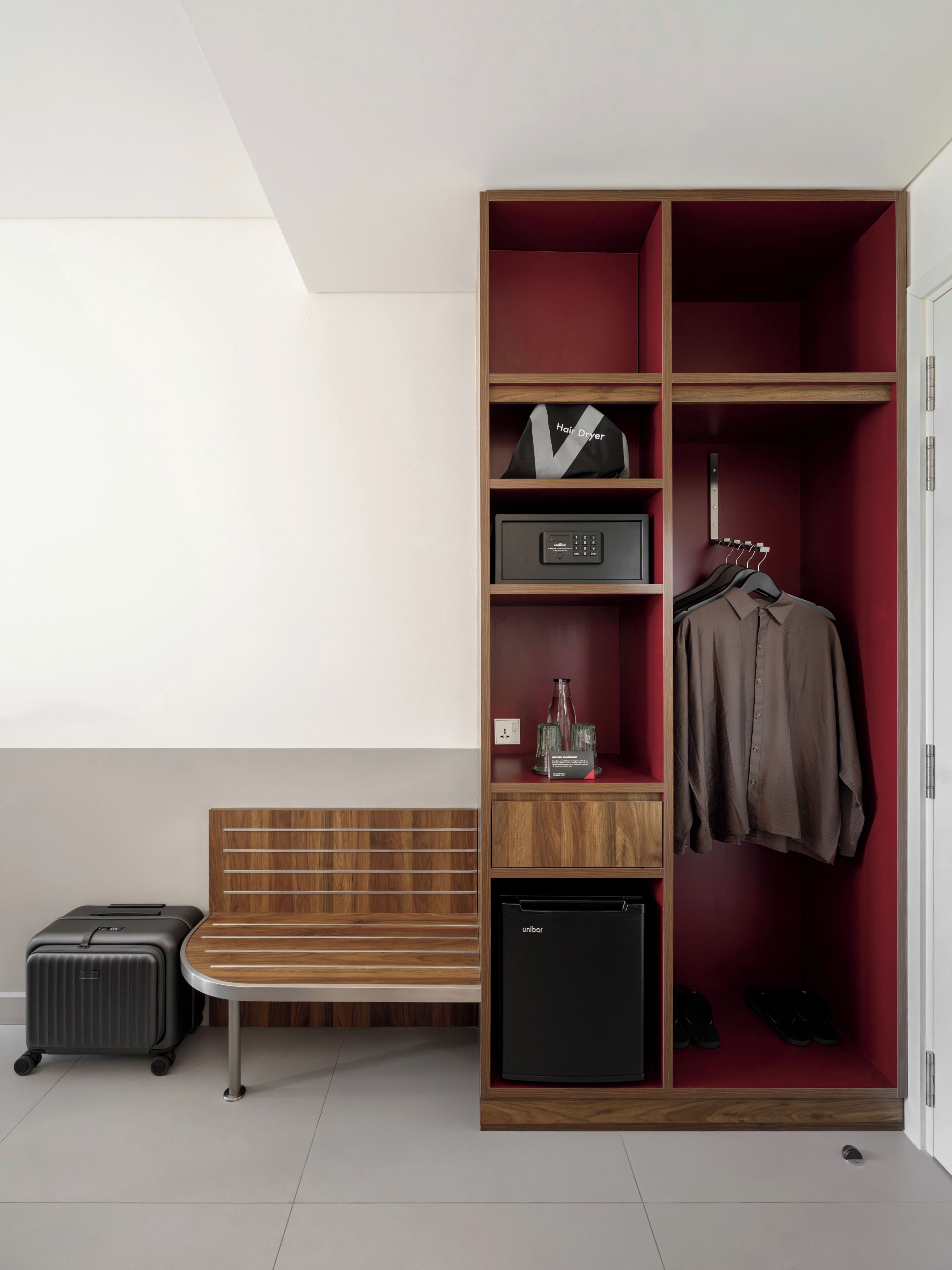 A hotel room with a bench, a wardrobe with hanging clothes, a hair dryer, and a small appliance.