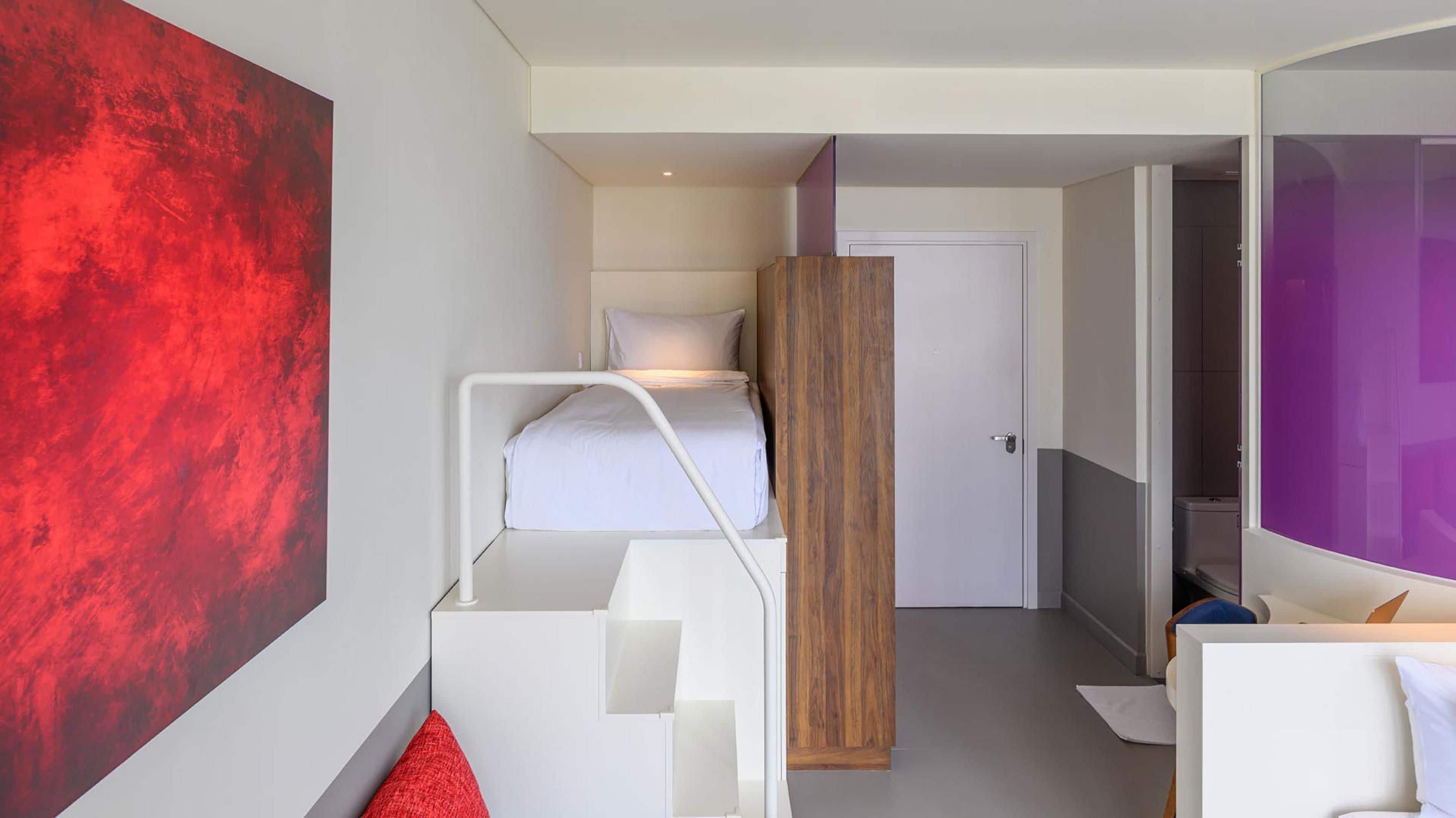 Une petite pièce comprenant un lit superposé, une armoire en bois, une porte donnant sur une autre pièce et un grand miroir.