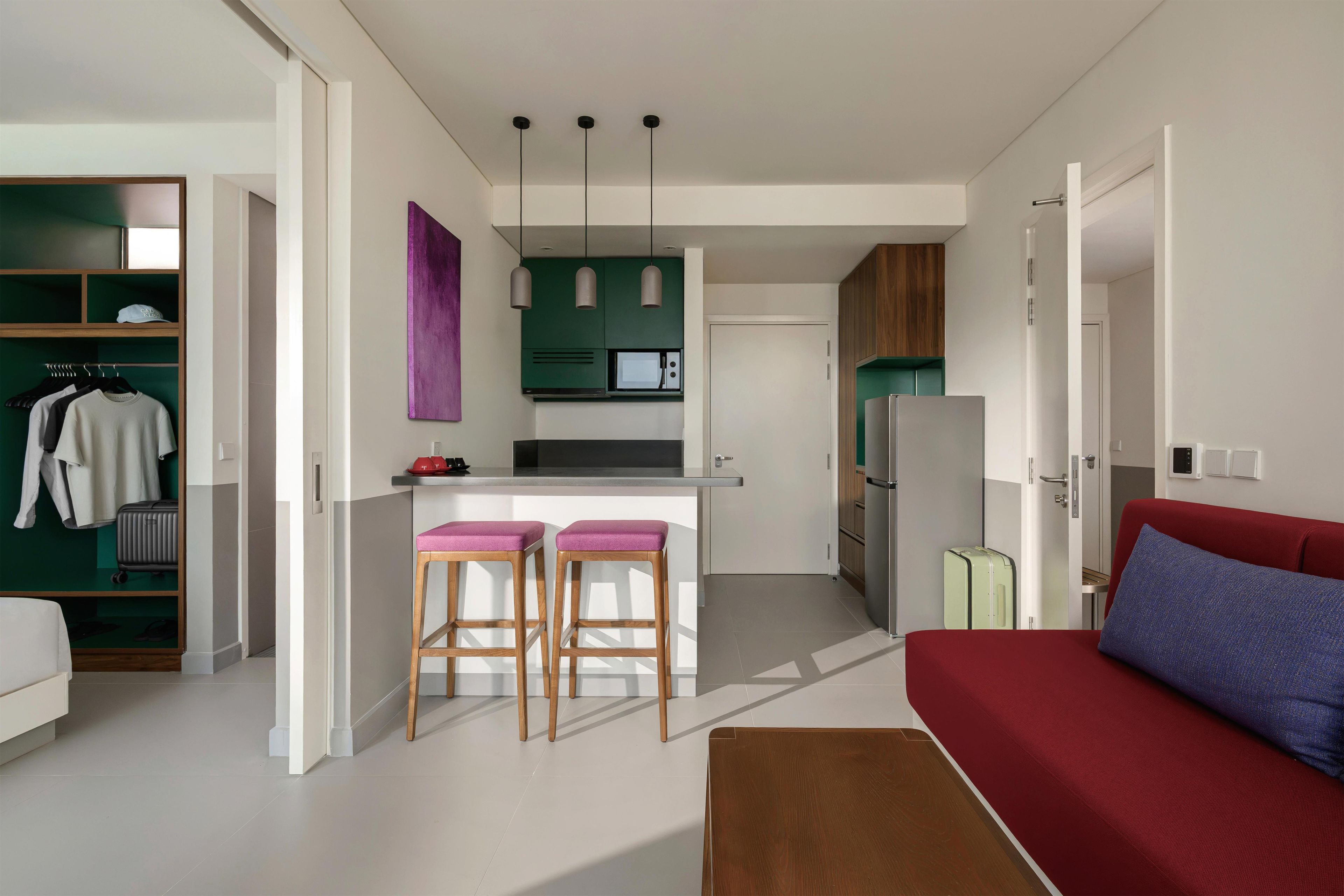 Een compacte leefruimte met een kitchenette, een eethoek met twee barkrukken, een rode bank en een kleine tafel.