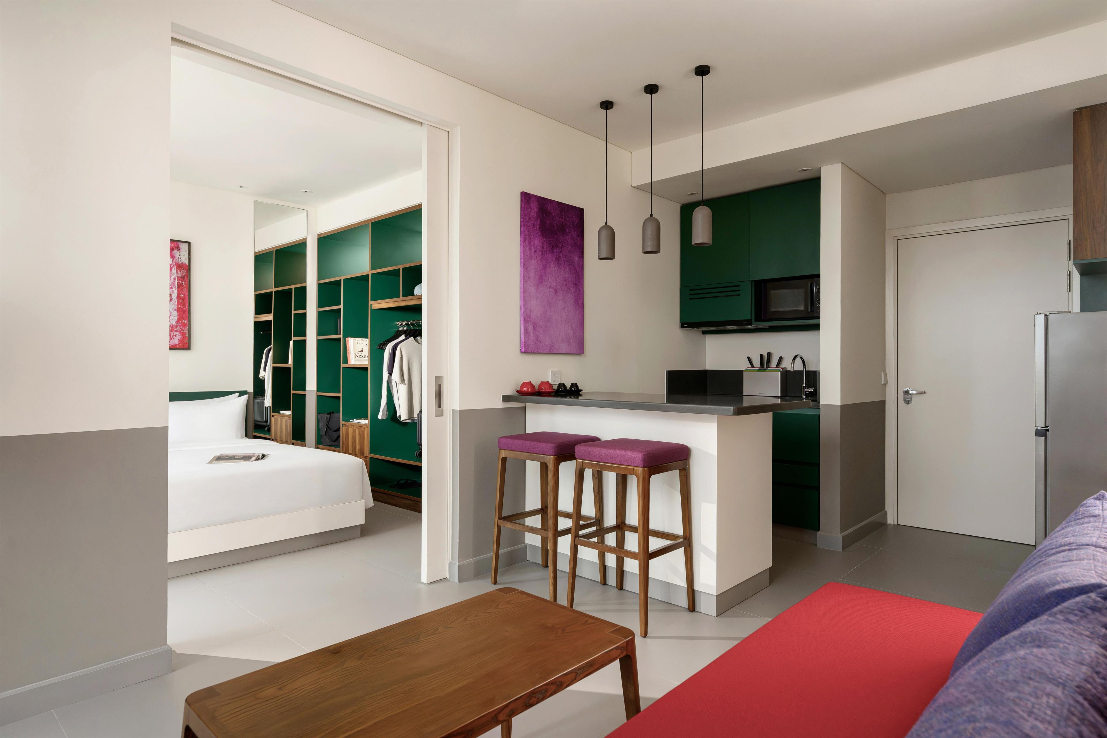 A modern hotel room with a bed, a wooden table, a kitchen area with a microwave, and a seating area with stools.