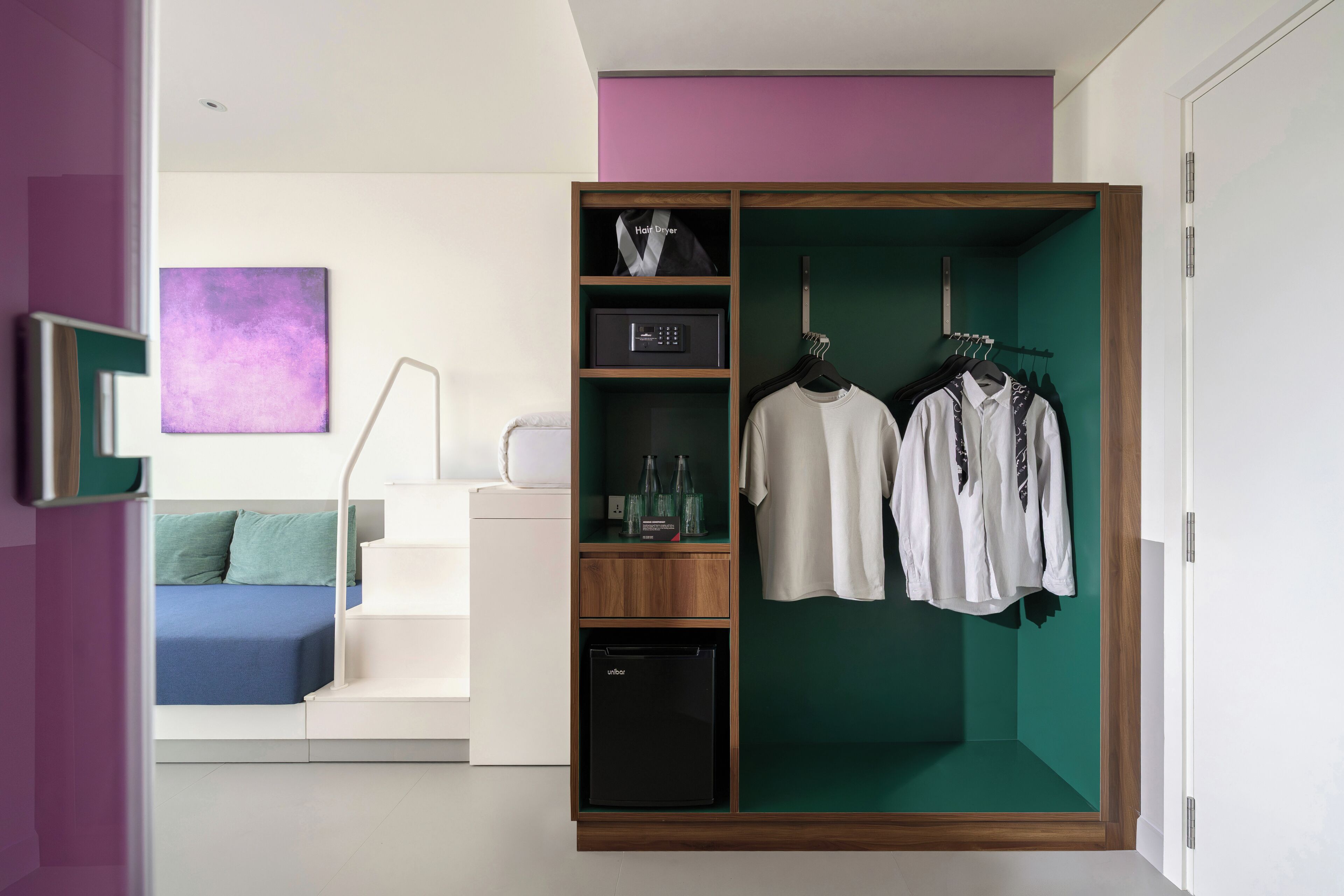 A modern bedroom with a walk-in closet, a bed with a blue headboard, and a purple abstract painting.