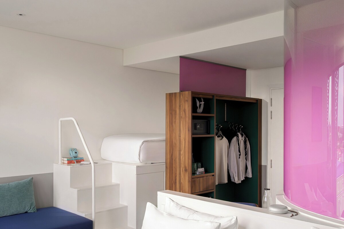 A modern bedroom with a white bed, a blue sofa, a wooden wardrobe, and a pink accent wall.