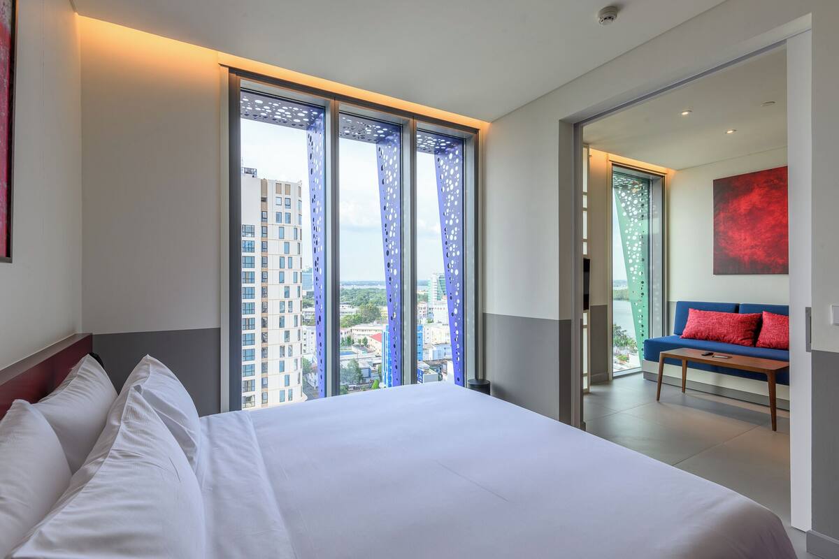 Habitación de hotel con una cama grande, vistas de la ciudad a través de ventanales del suelo al techo, un sofá azul con cojines rojos y un cuadro abstracto moderno en la pared.
