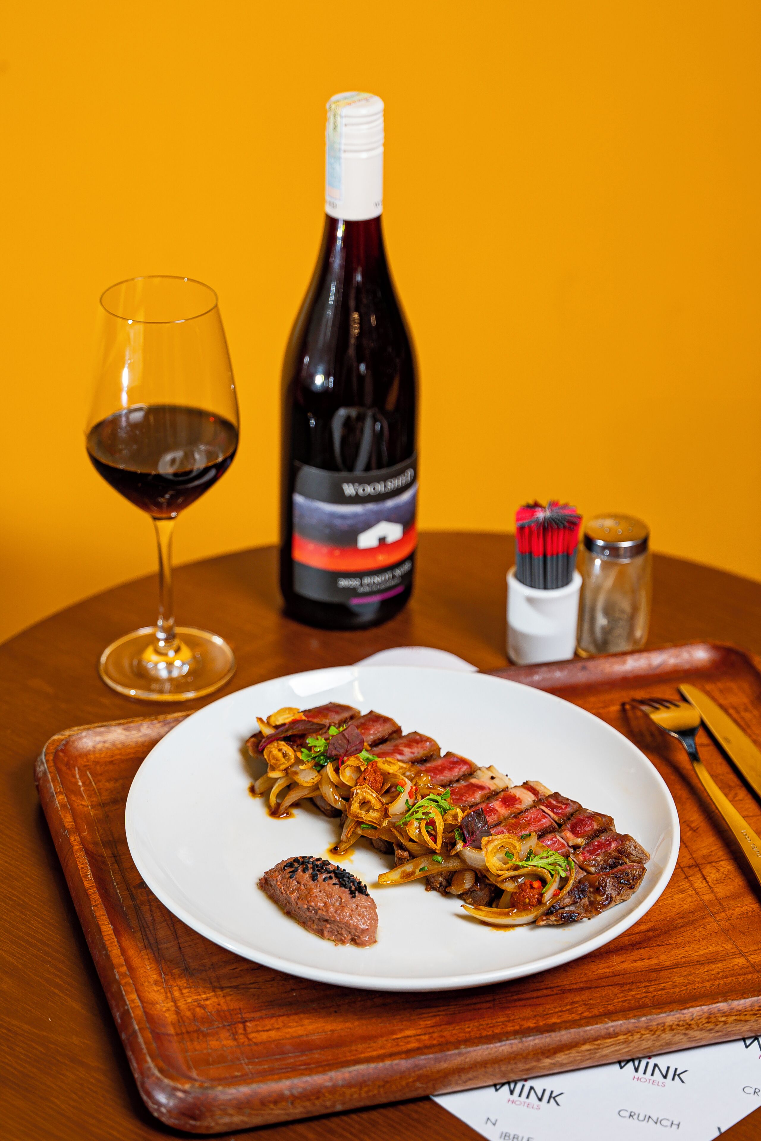 A plate of steak with onions, a glass of red wine, and a bottle of wine on a wooden table.
