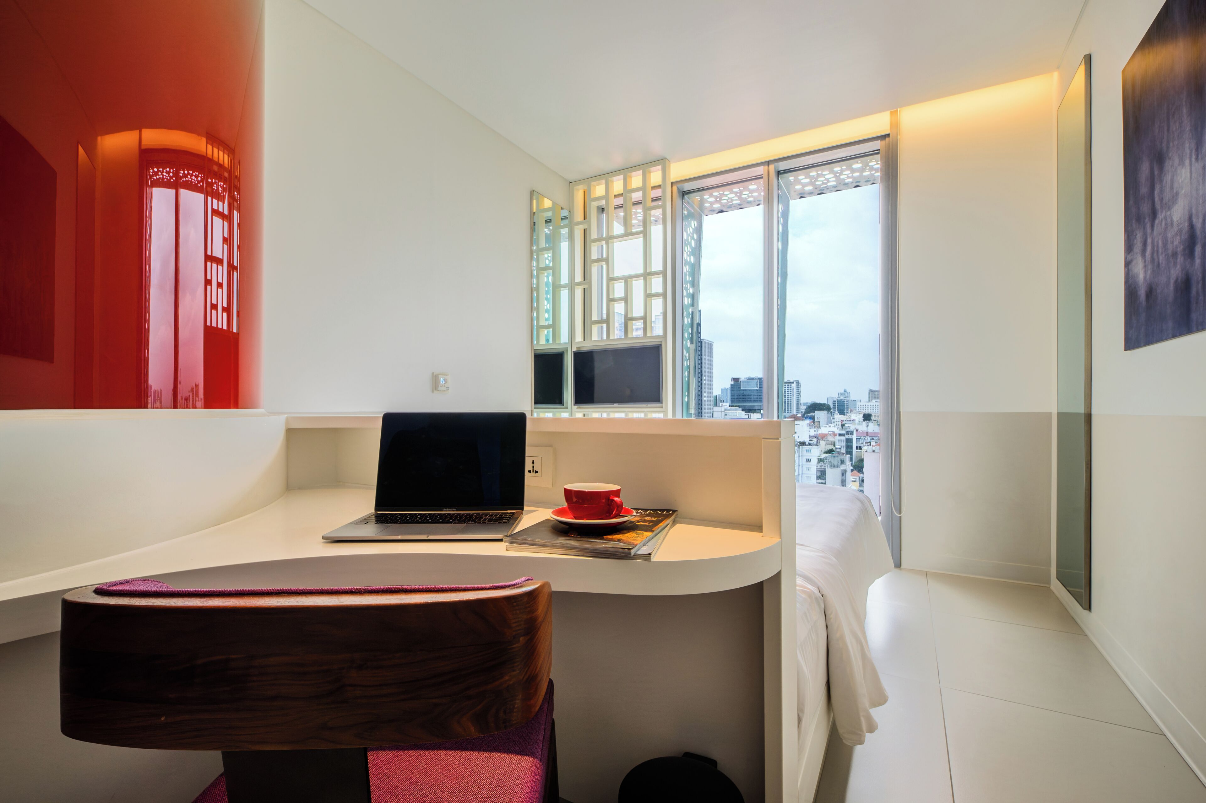 Una habitación moderna con un ordenador portátil sobre un escritorio, una taza roja y vistas de la ciudad a través de amplias ventanas.
