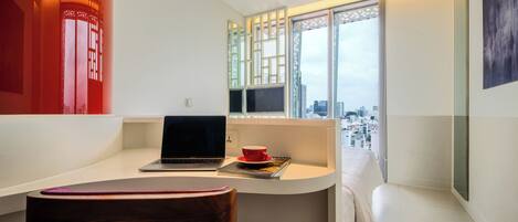 Una habitación moderna con un ordenador portátil sobre un escritorio, una taza roja y vistas de la ciudad a través de amplias ventanas.