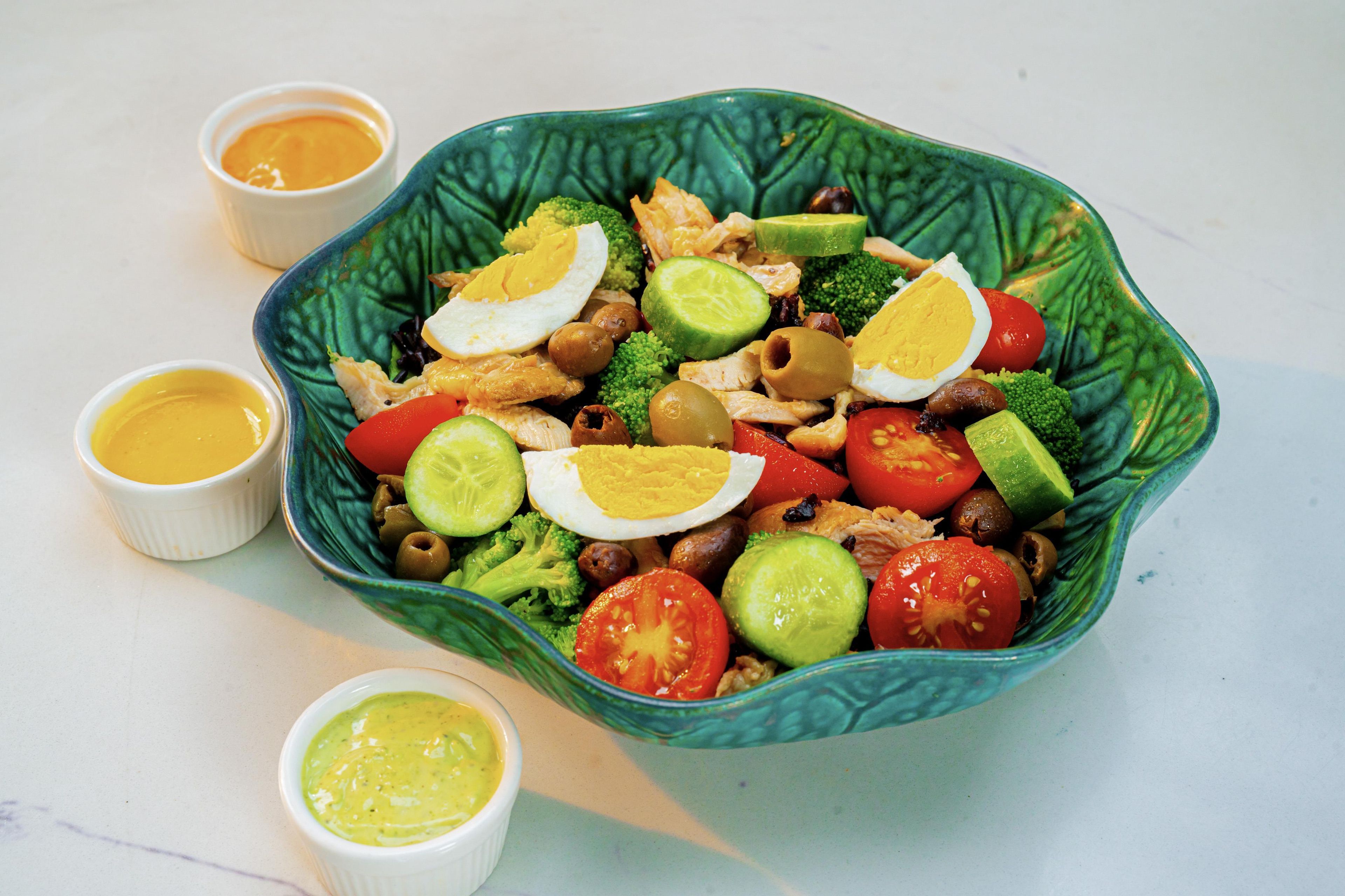 A bowl of salad with various vegetables and eggs, accompanied by two small bowls of dressing.