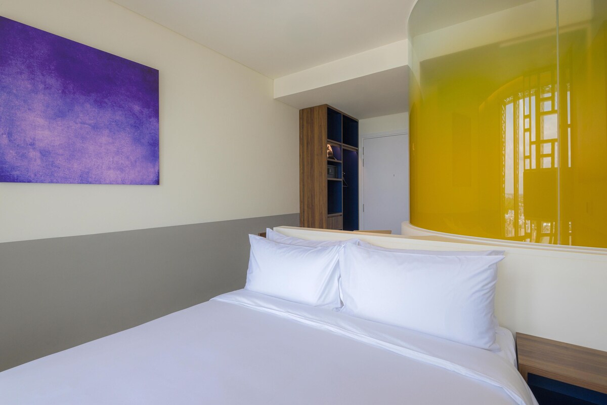 Une chambre d’hôtel avec un lit, une œuvre d’art violette accrochée au mur et une grande fenêtre qui reflète une lumière jaune.