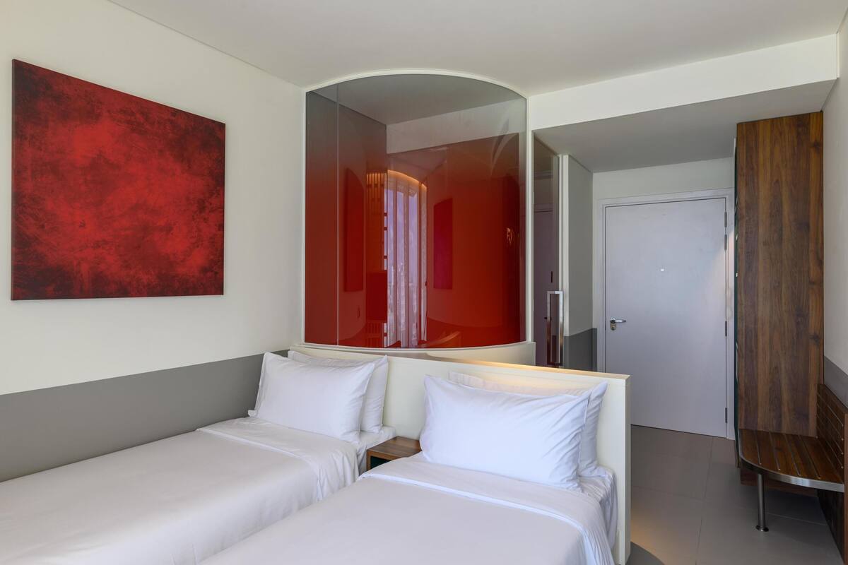 Habitación de hotel con dos camas, un espejo grande, un cuadro abstracto rojo y un banco de madera.
