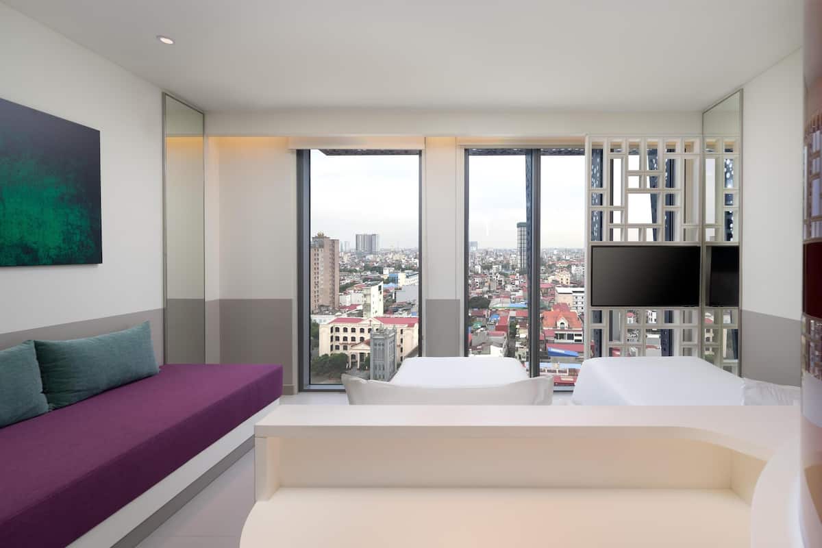 Una habitación de hotel moderna con un ventanal grande, un sofá, una cama, un televisor y una estantería.