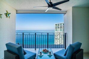A balcony with a view of the ocean, a table with pastries and coffee, and a ceiling fan.