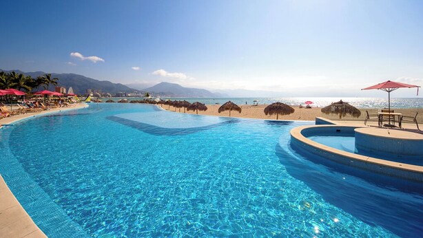 A clear blue pool with a curved edge, surrounded by beach umbrellas and lounge chairs, with a mountain view in the background.