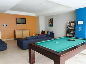 A pool table with a white ball, a sofa set, a bookshelf, and a television on the wall. - Oceanfront 22nd floor with 5 pools, Hotel Zone (Puerto Vallarta)
