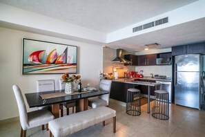 A modern kitchen with a dining area, featuring a large painting, a stainless steel refrigerator, and a coffee machine.