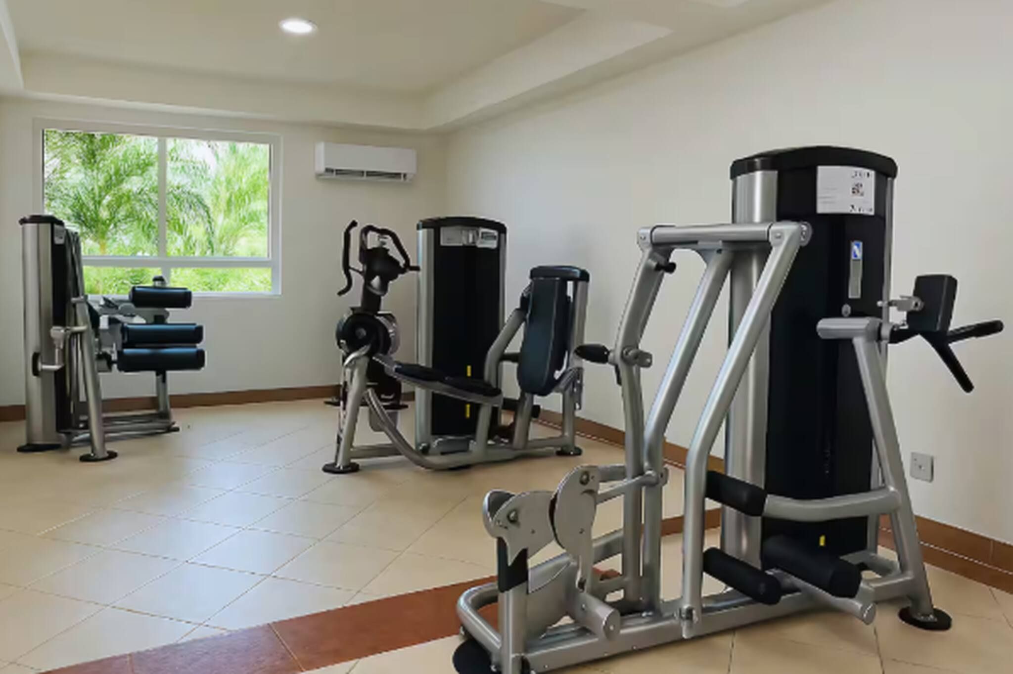 Un gimnasio con varias máquinas de ejercicio, ventanas amplias y piso de baldosas.