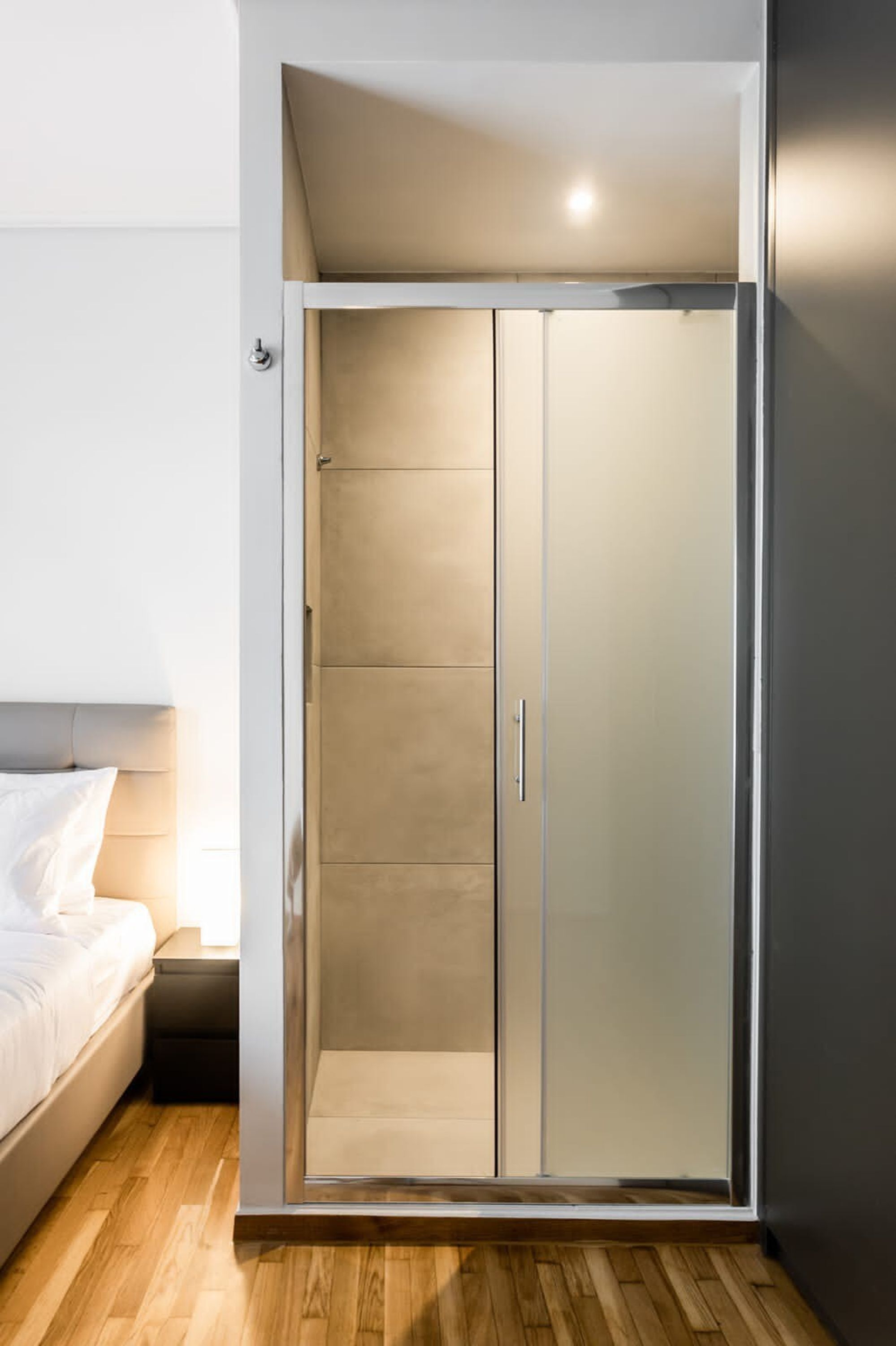 Una habitación moderna con un armario y puerta corredera de cristal, una cama con ropa de cama blanca y suelo de madera.