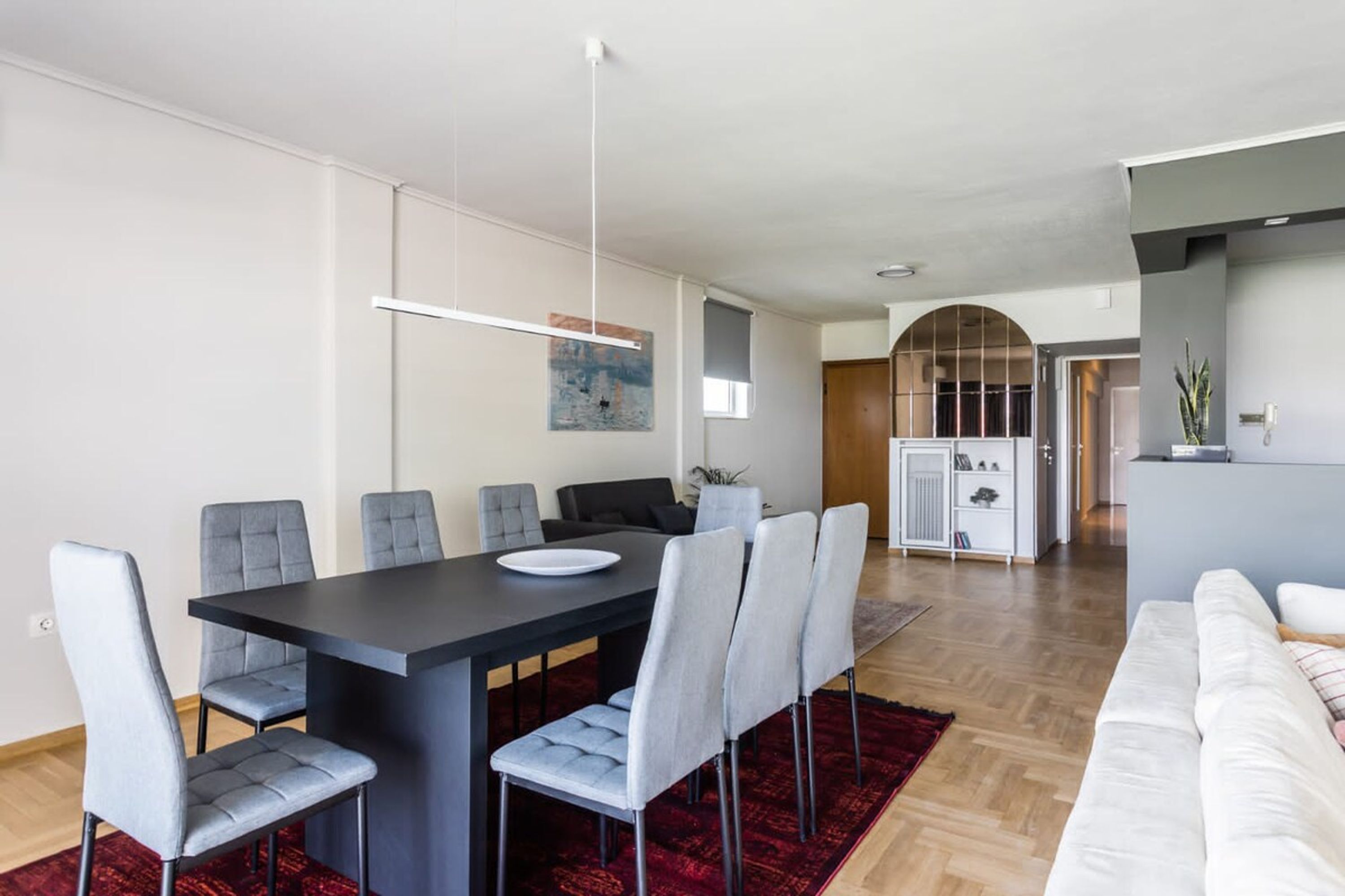 Una sala de estar moderna con comedor, que incluye una mesa grande, sillas tapizadas de gris, un tapete rojo y un sofá blanco.