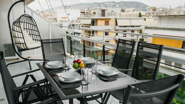 A balcony with a dining table set for four, surrounded by chairs, with a hanging chair and a view of the cityscape. - Spacious Seaside Flat at Alimos (Alimos)