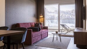 A modern living room with a sofa, chairs, a small table, and a balcony with a view of snow-covered mountains.