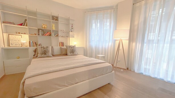Une chambre spacieuse avec un grand lit, une table de chevet, une lampe sur pied et une bibliothèque remplie de livres et d’objets décoratifs.