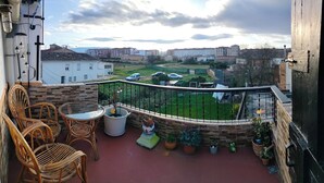 Un balcon agrémenté de chaises et d’une table en osier, entouré de plantes en pot et offrant une vue sur un quartier résidentiel avec ses immeubles et son espace vert.